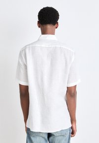 Camisa de lino blanca de manga corta con cuello, detalles de costura visibles y un corte relajado, mostrada desde la parte posterior sobre un fondo claro.