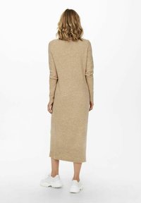 Robe beige en maille jusqu'au genou avec manches longues et coupe droite, associée à des baskets blanches. Texture douce avec des détails côtelés à l'ourlet.