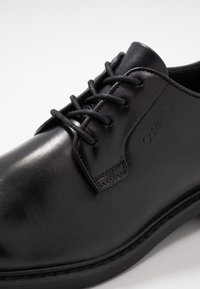 Chaussure de ville en cuir noir avec une finition lisse, dotée de lacets ronds et d'un logo en relief sur le côté. Détails cousus et semelle robuste.