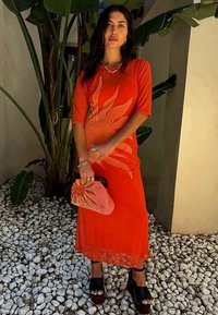 Rød kjole med bladmønster, knelange, myk stoff. Svart platåsandaler, holder en rosa clutch-veske. Grønt løvverk og småstein i bakgrunnen.