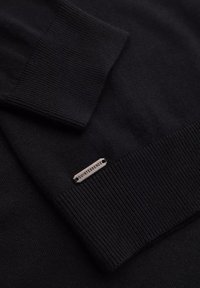 Svart stickad tröja med ribbade ärmslut och en metalltagg som står "QUINTESSENCE." Slät textur med en enhetlig yta.