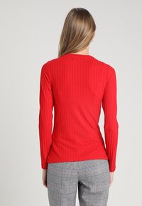 Donna con capelli lunghi che indossa un top rosso a coste con maniche lunghe e pantaloni a quadretti grigi, vista di spalle su uno sfondo neutro.