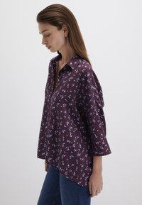 Dark purple button-up shirt featuring light blue floral print, rolled sleeves, and a curved hem, made from soft fabric.