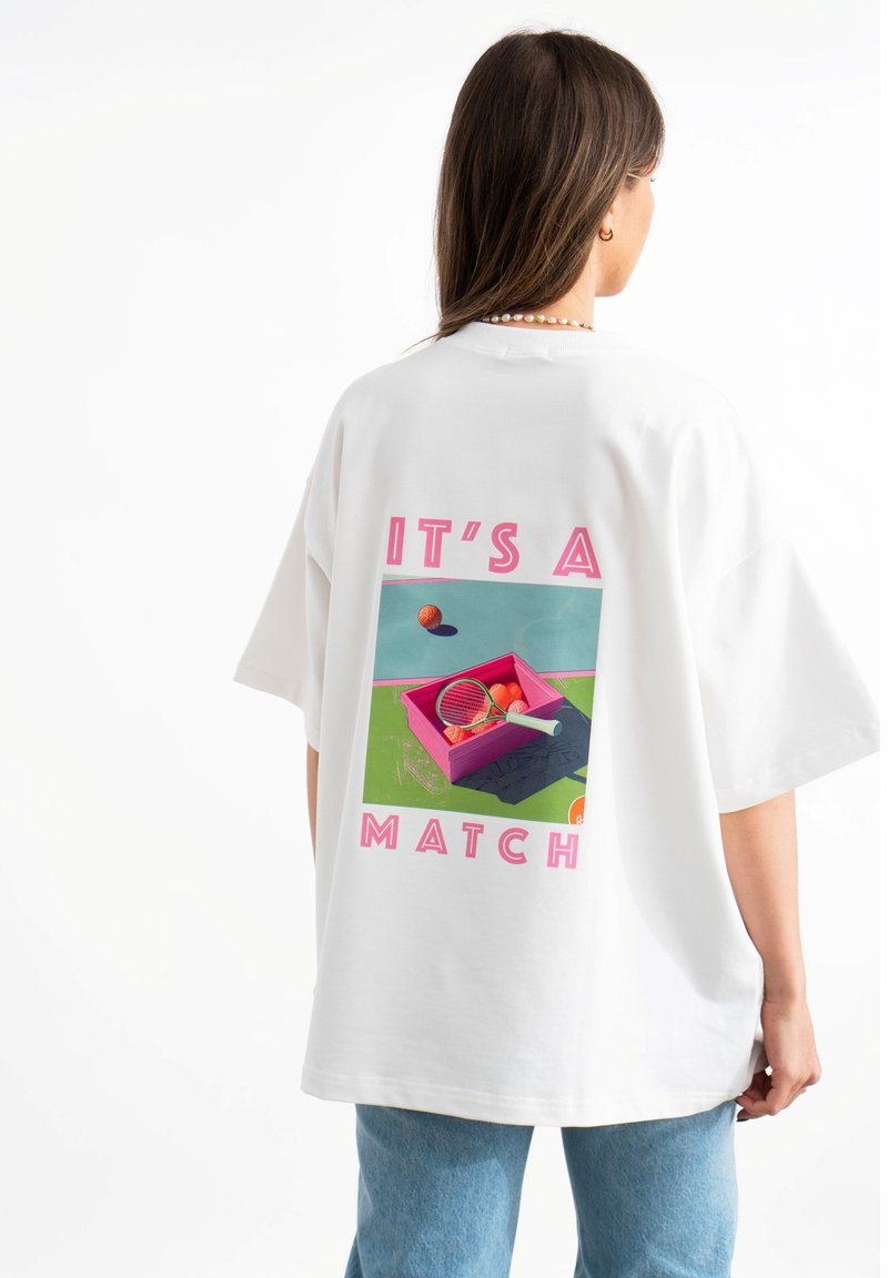 Biała, oversize'owa koszulka z kolorową grafiką na plecach, z napisem "TO DOPASOWANIE" oraz różowym pudełkiem zawierającym sprzęt tenisowy.