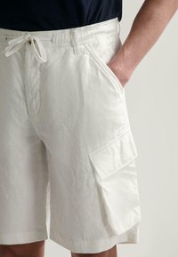 Weiße Leinen-Cargo-Shorts mit elastischem Bund und Kordelzug, ausgestattet mit seitlichen Taschen und einer Klapptasche am Bein zur Aufbewahrung.