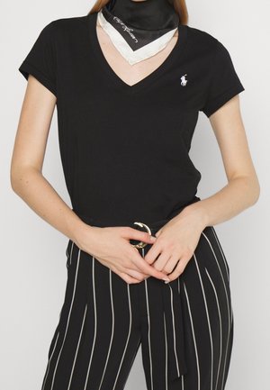 Femme portant un t-shirt noir à col en V avec un petit logo blanc, une écharpe noire et blanche, et un pantalon noir taille haute avec des rayures verticales blanches et une ceinture.