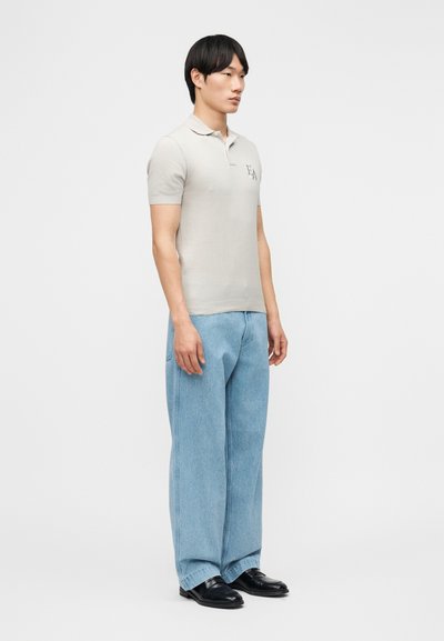 Άνδρας που στέκεται πλευρικά φορώντας ανοιχτό γκρι κοντομάνικη πόλο μπλούζα, φαρδιά ανοιχτά μπλε τζιν και μαύρα παπούτσια σε λευκό φόντο.