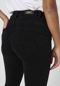Gros plan d'une personne portant un jean noir taille haute avec des poches arrière et une étiquette "ONLY", associé à une chemise blanche rentrée.