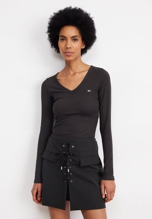 Zwarte ribbel top met lange mouwen en een v-hals, voorzien van een logopatch, gecombineerd met een zwarte rok met een frontslit en een vetersluiting.