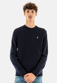 Maglione girocollo blu navy realizzato in morbido tessuto a maglia, con un piccolo logo a forma di albero rosso sul petto e polsini e orlo a coste.