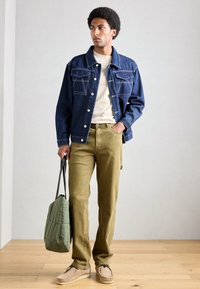 Denimjakke med kontraststing, creme T-shirt, olivengrønne bukser, tan-farvede fodtøj og en quiltet grøn tote bag, sat mod en neutral baggrund.