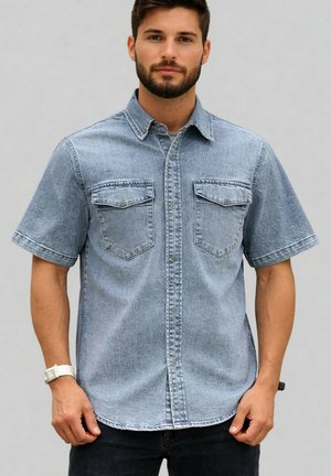Jeune homme avec une barbe courte portant une chemise en jean bleu clair à manches courtes avec des poches avant, sur un fond uni.