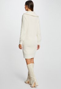 Abito a maglia bianco e morbido con colletto a scialle, maniche lunghe e silhouette aderente, abbinato a stivali beige al ginocchio con tacco a blocco.