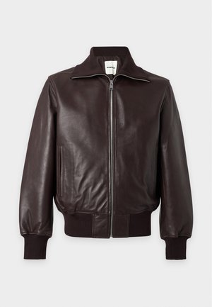 Jeune homme debout, les mains dans les poches, portant une veste en cuir marron foncé, une chemise noire, un pantalon large bleu marine et des chaussures noires sur fond blanc.
