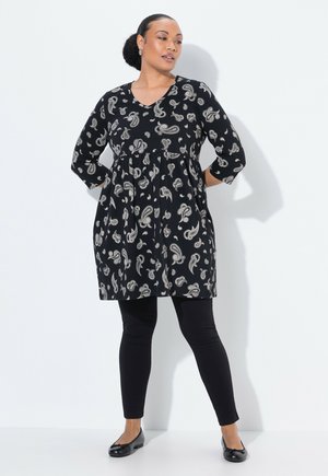 Schwarzes Tunika-Kleid mit Paisleymuster, Dreiviertelärmel und V-Ausschnitt, kombiniert mit schwarzen Leggings und flachen Schuhen. Glattes Material, entspannter Schnitt.
