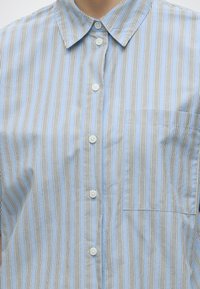 Gros plan d'une chemise bleu clair avec des rayures verticales blanches et noires, des boutons blancs, un col et une poche poitrine sur le côté droit.
