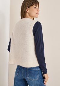 Beige, lurvig stickad väst med rund halsringning och ärmlös design, bärs över en marinblå långärmad topp. Har en kortad längd och ribbad fåll.