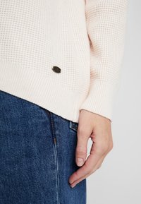 Suéter de punto de color rosa claro con un patrón texturizado y puños acanalados, que presenta un pequeño detalle de logotipo metálico oscuro en el dobladillo. Llevado sobre un vaquero.