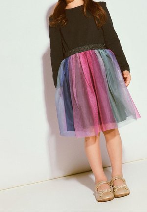 Enfant portant un haut noir à manches longues et une jupe en tulle colorée aux nuances de rose, violet et bleu, assortie de chaussures Mary Jane dorées.