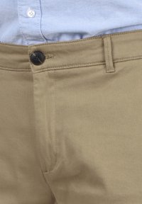 Pantalones caqui con una textura suave, con un cierre de botón negro, trabillas para cinturón y costuras limpias, parcialmente mostrados con una camisa de color azul claro.