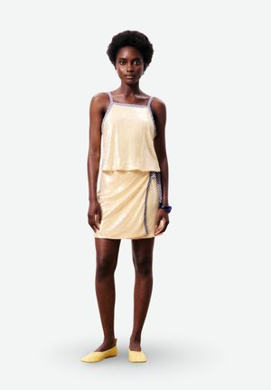 Femme portant une robe sans manches de couleur crème avec un bord à carreaux et des ballerines jaunes, debout devant un fond blanc uni.