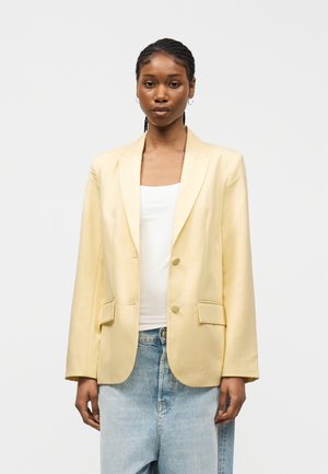 Młoda kobieta z zaplecionymi włosami, ubrana w jasny żółty żakiet, biały top i luźne, jasnoniebieskie dżinsy, stoi na jednolitym tle.