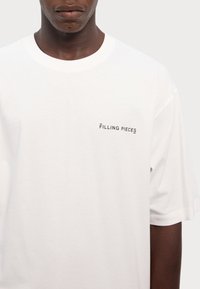 Hvid bomulds-t-shirt med rund hals og korte ærmer. Et lille sort logo, "FILLING PIECES," er trykt på venstre bryst.