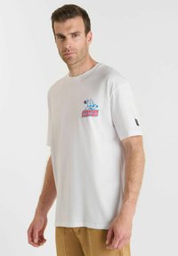 Man in een wit T-shirt met een kleine afbeelding en de tekst "LET OP BOZE BAAS" op de borst, gecombineerd met lichtbruine broek.