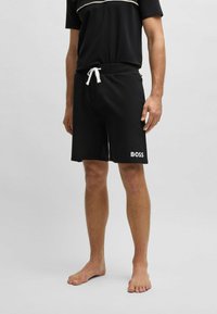 Shorts neri con cintura elasticizzata e laccetto bianco. Presentano un logo bianco "BOSS" sulla gamba sinistra. Texture del tessuto liscia.