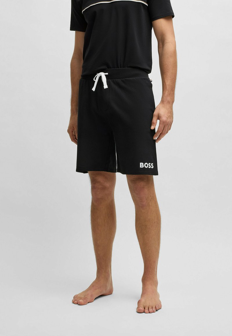 Shorts neri con cintura elasticizzata e laccetto bianco. Presentano un logo bianco "BOSS" sulla gamba sinistra. Texture del tessuto liscia.
