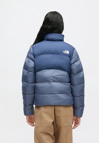 Hosszú sötét hajú személy, aki kék és világoskék North Face pufinakabátot és bézs nadrágot visel, háttal áll egy sima háttér előtt.