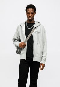 Νέος άντρας που φοράει ανοιχτό γκρι ζακέτα με φερμουάρ Calvin Klein, μαύρο πουκάμισο, μαύρο παντελόνι, ασημένιο κολιέ με αλυσίδα και λουράκι τσάντας πάνω από τον ώμο.