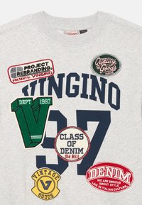 Pelēks džemperis, uz kura ir dažādi krāsaini auduma pielikumi, tostarp teksti un grafikas: "VINGINO," "CLASS OF DENIM," un "VINTAGE GOODS."