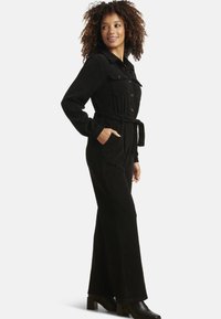 Tuta in velluto nero con vita aderente, maniche lunghe, tasche sul petto e cintura. Abbinata a stivali neri con tacco alto.