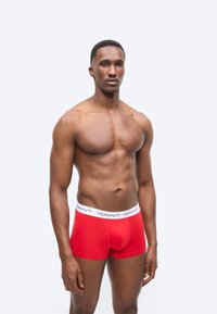 Μυώδης άνδρας που στέκεται φορώντας κόκκινα μπόξερ με λευκή μέση με την ένδειξη "GANT" σε απλό φωτεινό φόντο.