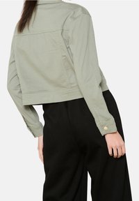 Kobieta w krótkiej, jasnozielonej kurtce z zapiętymi mankietami i czarnych spodniach z wysokim stanem, pokazana z tyłu na wysokości talii.