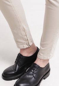 Pantalon beige avec fermetures éclair à la cheville porté avec des chaussures en cuir noires à lacets sur fond blanc.