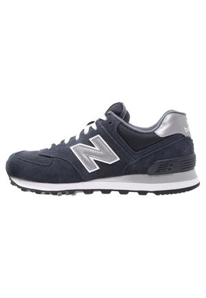 Dunkelgrauer New Balance Sneaker mit weißen Schnürsenkeln, silbernem Logo, Mesh- und Wildlederobermaterial und Gummisohle, seitlich betrachtet.