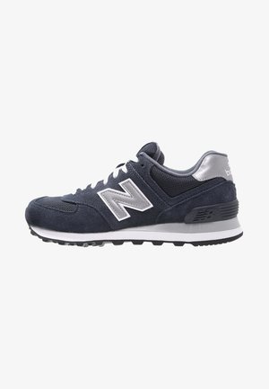 Ciemnoszary sneaker marki New Balance z białymi sznurówkami, srebrnym logo, siateczkową i zamszową cholewką oraz gumową podeszwą widoczny z boku.