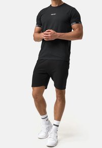 Czarny strój sportowy składający się z t-shirtu z krótkim rękawem z logo oraz dopasowanych szortów. Uzupełniony białymi sneakersami i pasiastymi skarpetami do kostki.