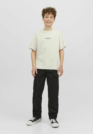 Ljusbeige bomullströja med korta ärmar och en logo på bröstet, matchad med svarta cargobyxor och svarta sneakers med vita sulor.