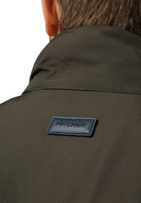 Oliwkowa kurtka z wysokim kołnierzem i niebieską skórzaną naszywką z wytłoczonym tekstem "PRÓCHNIK" z tyłu. Gładka tekstura.