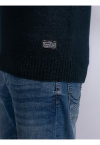 Maglione lavorato a maglia blu scuro con orlo a coste, con un'etichetta che recita "Petrol Ind" sul lato sinistro in basso, abbinato a jeans azzurri.