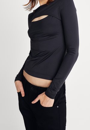 Kobieta ubrana w dopasowaną czarną bluzkę z długim rękawem z ukośnym wycięciem na piersi oraz czarne spodnie, ręce spoczywają w kieszeniach.