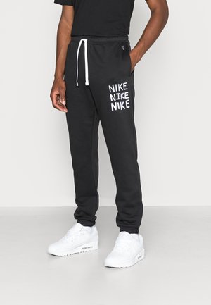 Osoba nosząca czarne spodnie dresowe Nike z białymi sznurkami i powtarzającymi się logo Nike, w połączeniu z białymi trampkami i czarną koszulką.