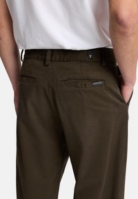Pantaloni di cotone marroni con una texture liscia, dotati di due tasche posteriori e di una piccola etichetta sulla vita. Abbinati a una camicia bianca.