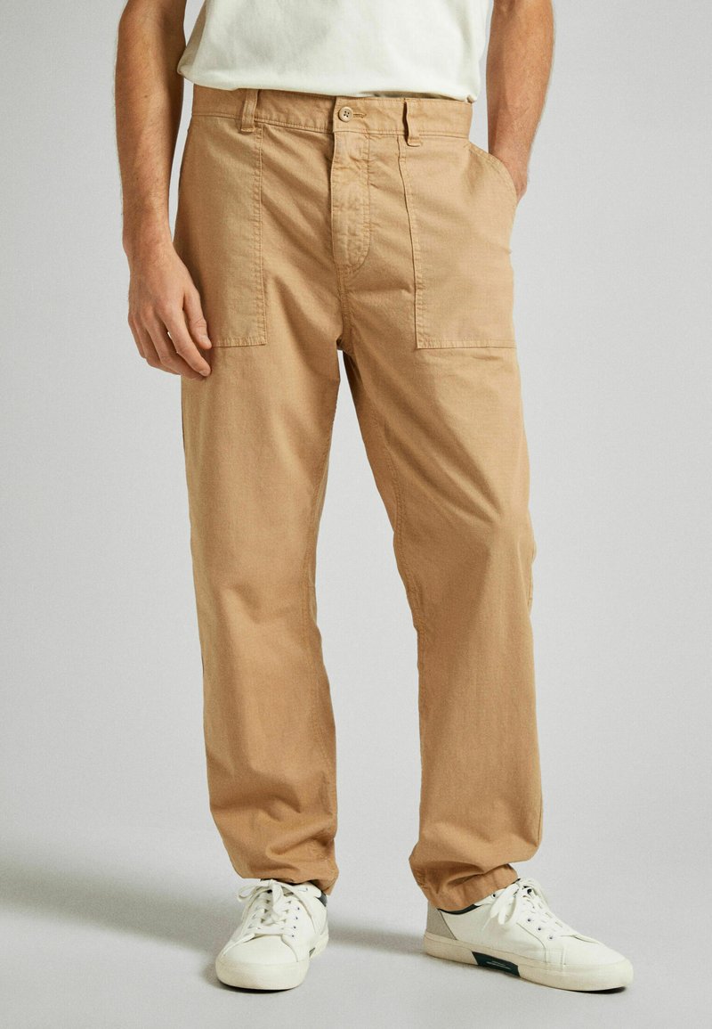 Pantalones de algodón color caqui con un corte relajado, que cuentan con dos bolsillos frontales y un cierre de botón, combinados con zapatillas blancas.