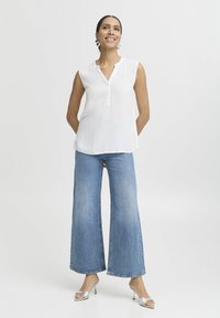 Weiße ärmellose Bluse mit Knopfleiste, kombiniert mit weit geschnittenen blauen Denim-Jeans und silbernen Absatzsandalen. Einfaches und modernes Design.