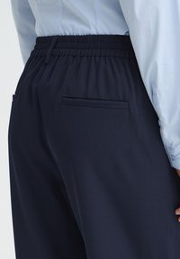 Pantalon bleu marine avec une ceinture élastique, doté d'une texture lisse et de deux poches arrière. Porté avec une chemise bleu clair  à boutons.