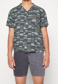 Camisa de manga corta con botones en patrones geométricos multicolores, con una paleta de colores en verde, gris y blanco. Combinada con pantalones cortos de mezclilla gris.